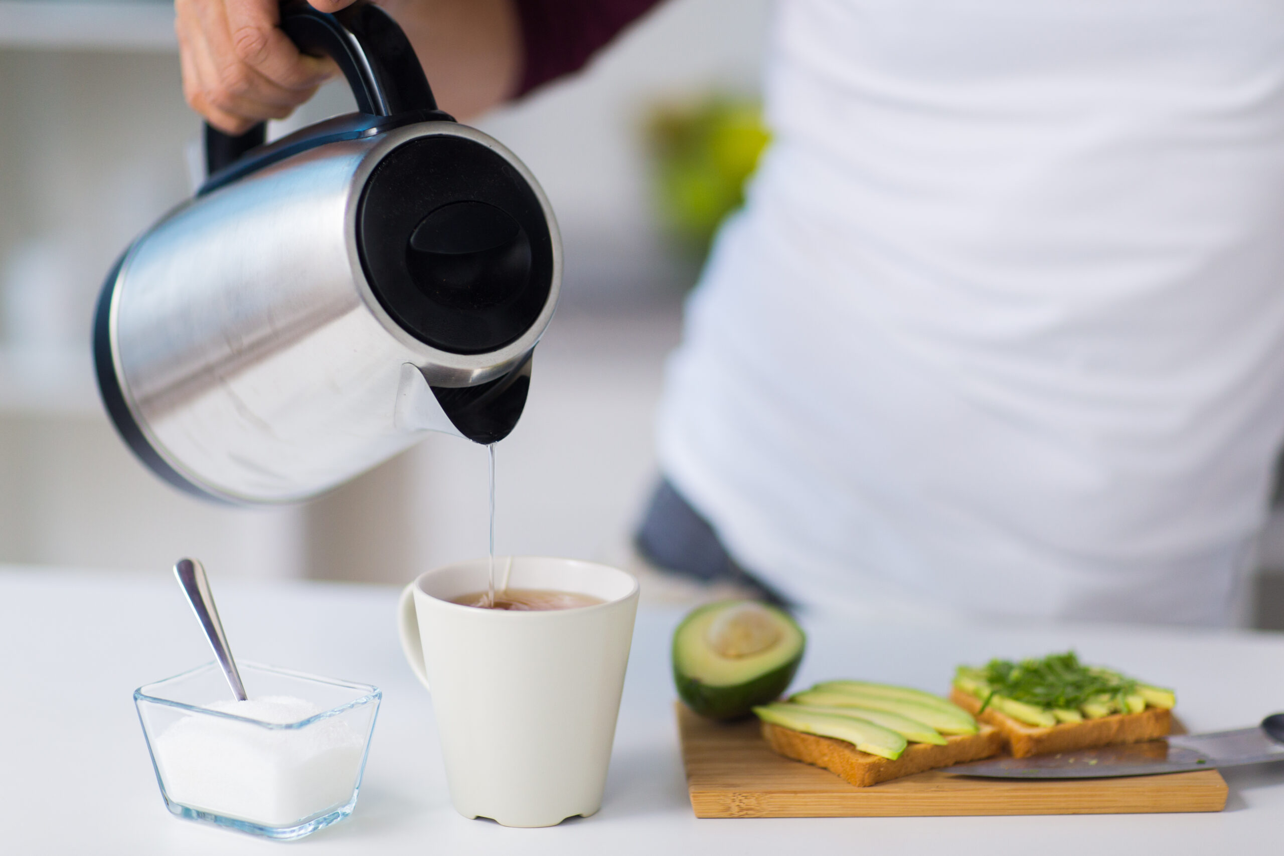 person pouring hot water from electric kettle into mug next to plate of avocado toast